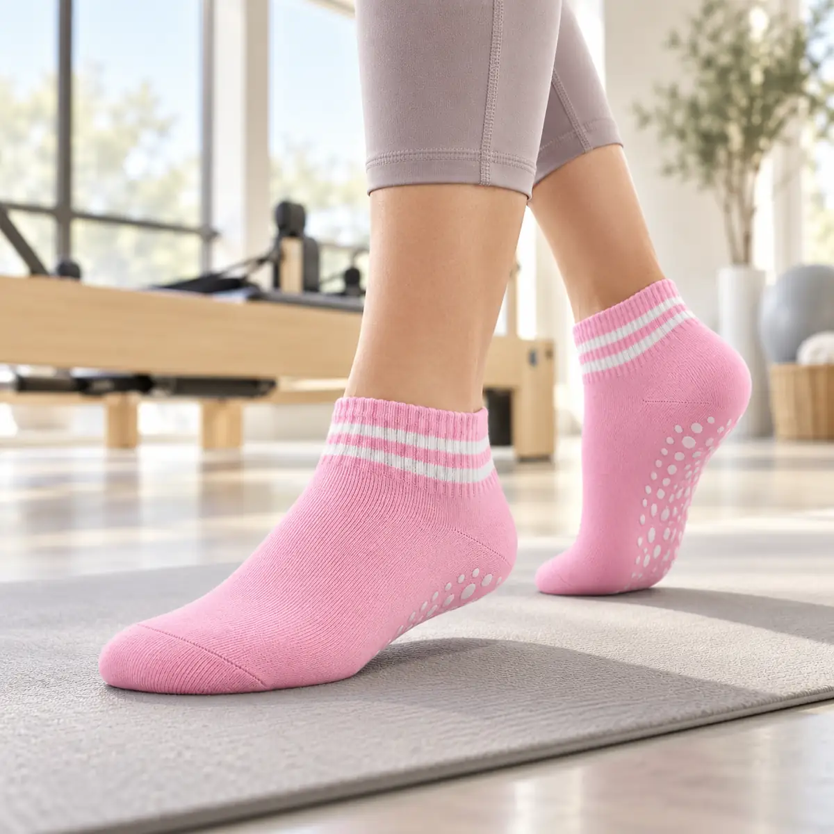 Roze Enkel sokken met twee witte strepen aan de voeten van een vrouw op een grijze mat, witte noppen onder de opgetilde hiel, lichte studio met een houten reformer onscherp op de achtergrond