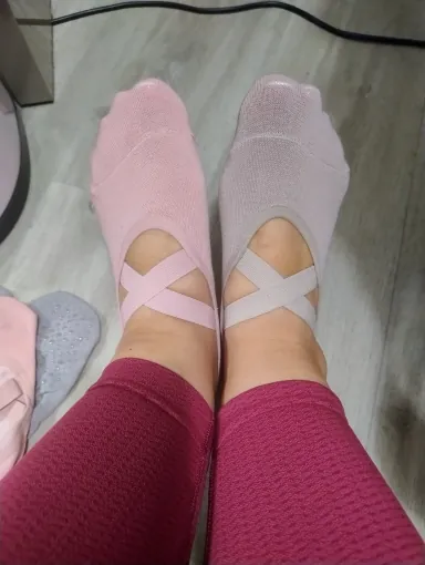 Twee voeten in roze balletsokken met gekruiste bandjes over de wreef, tenen bedekt, bordeauxrode legging; links op de vloer ligt een grijs paar.