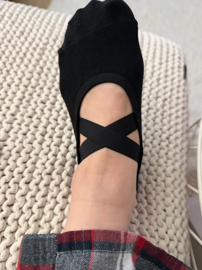 Eén voet in een zwarte balletsok met gekruiste bandjes over de open wreef, tenen bedekt, rustend op een grofgebreide crèmekleurige poef.